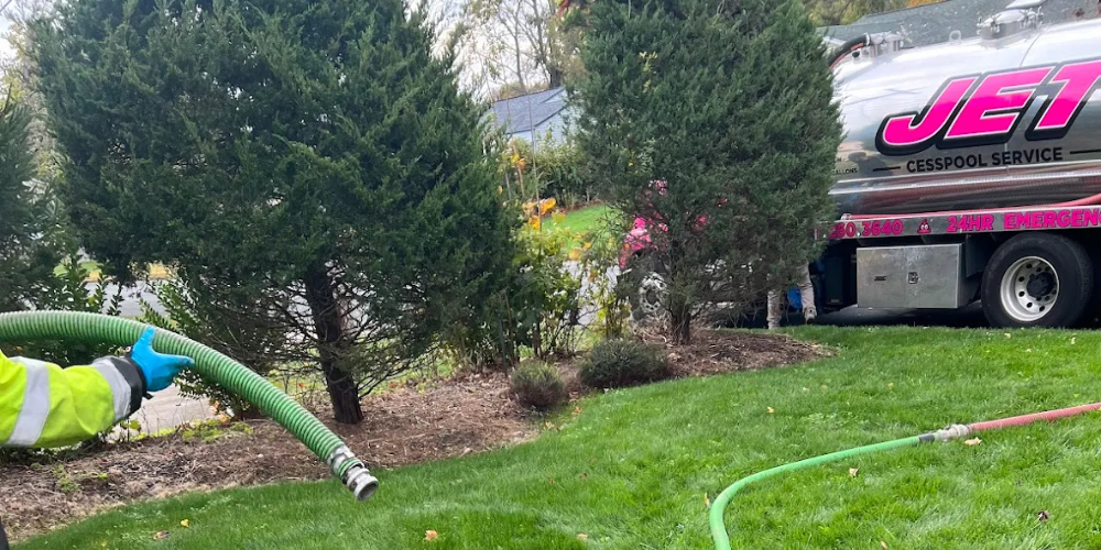 Cesspool pumping with worker holding green septic suction hose by Jet Cesspool Service truck near Betsy Ross Dr and Trailer Park, Bohemia, NY.