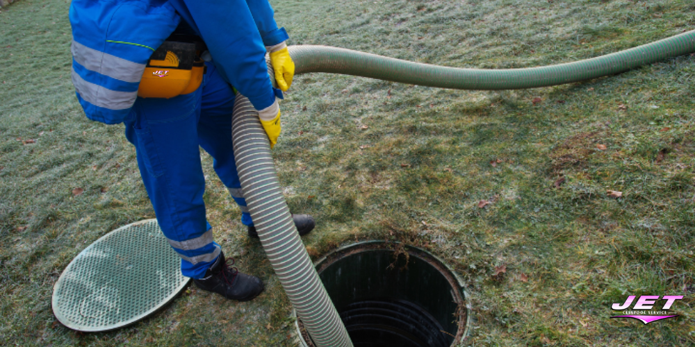 Cesspool pumping worker in blue safety gear holds a large suction hose into an open ground tank, green lid nearby, on frosty grass at Oak Forest Dr and Sampson Ave, Islandia, NY.