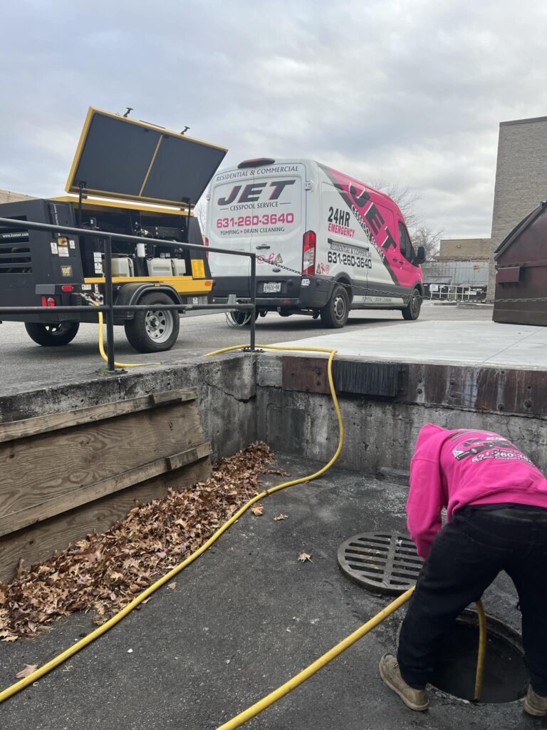 Cesspool pumping in Babylon, NY shows a worker in a pink Jet Cesspool Service sweatshirt holding a yellow suction hose tool beside an open septic cover, with the company van in view. Pamequa Rd and Annuskemunnica Rd, Babylon, NY.