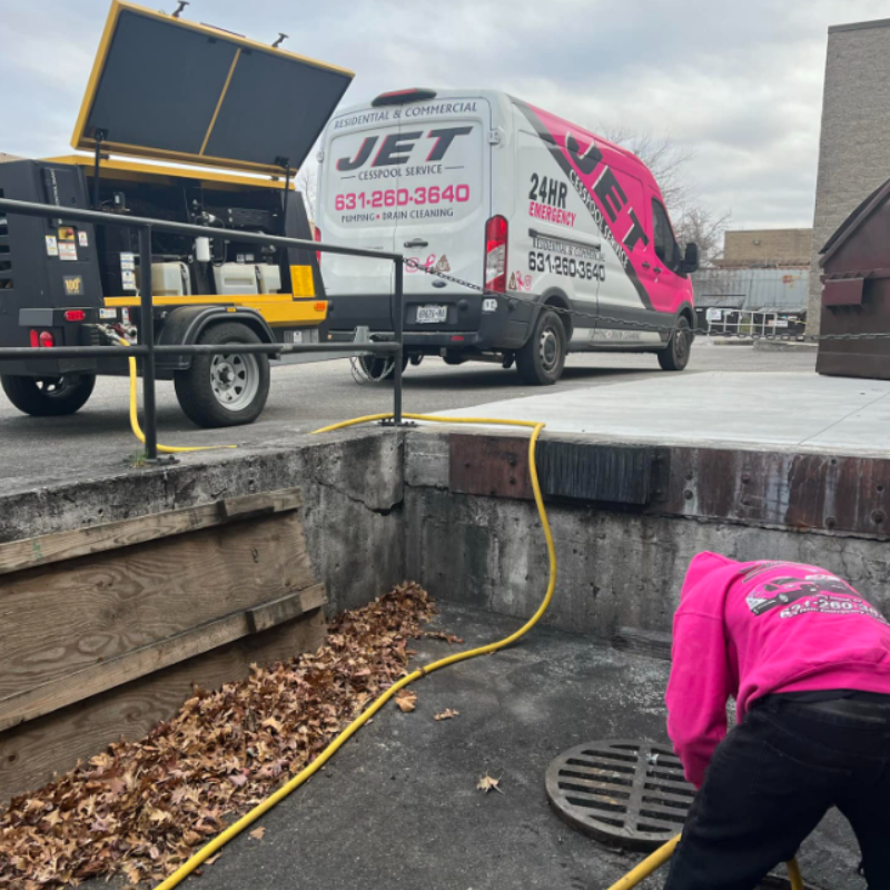 Drain cleaning in progress as a Jet Cesspool Service technician in a pink hoodie bends over a storm drain using a yellow hose. A branded service van and equipment trailer are parked nearby. The scene takes place outdoors near fallen leaves at Fire Island Ave and Prospect St, Babylon, New York.