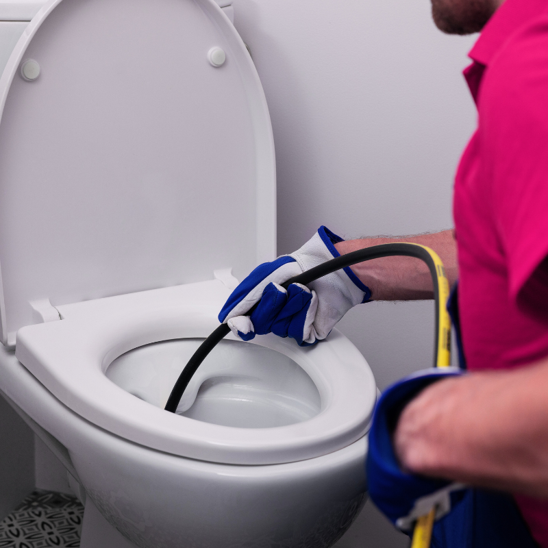 Clogged toilet repair shown as a worker in gloves inserts a flexible plumbing snake into a toilet bowl, gripping the auger cable while clearing a blockage during a hands-on service call in Long Island, NY.